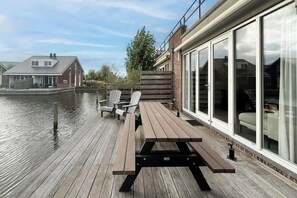 Una terraza de madera con mesas y sillas de picnic junto a una masa de agua, grandes puertas correderas de vidrio y un edificio de ladrillo al fondo.