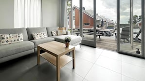 Een moderne woonkamer met een grijze bank, een houten salontafel en een grote schuifpui die uitkomt op een balkon met tuinmeubilair.