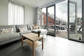 Een moderne woonkamer met een grijze bank, een houten salontafel en een grote schuifpui die uitkomt op een balkon met tuinmeubilair.