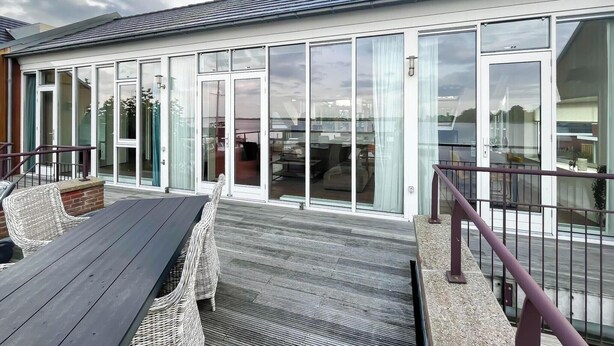A modern building with large glass doors, a wooden deck, wicker furniture, and a metal railing. - Penthouse with Lake View Terrace (Uitgeest)