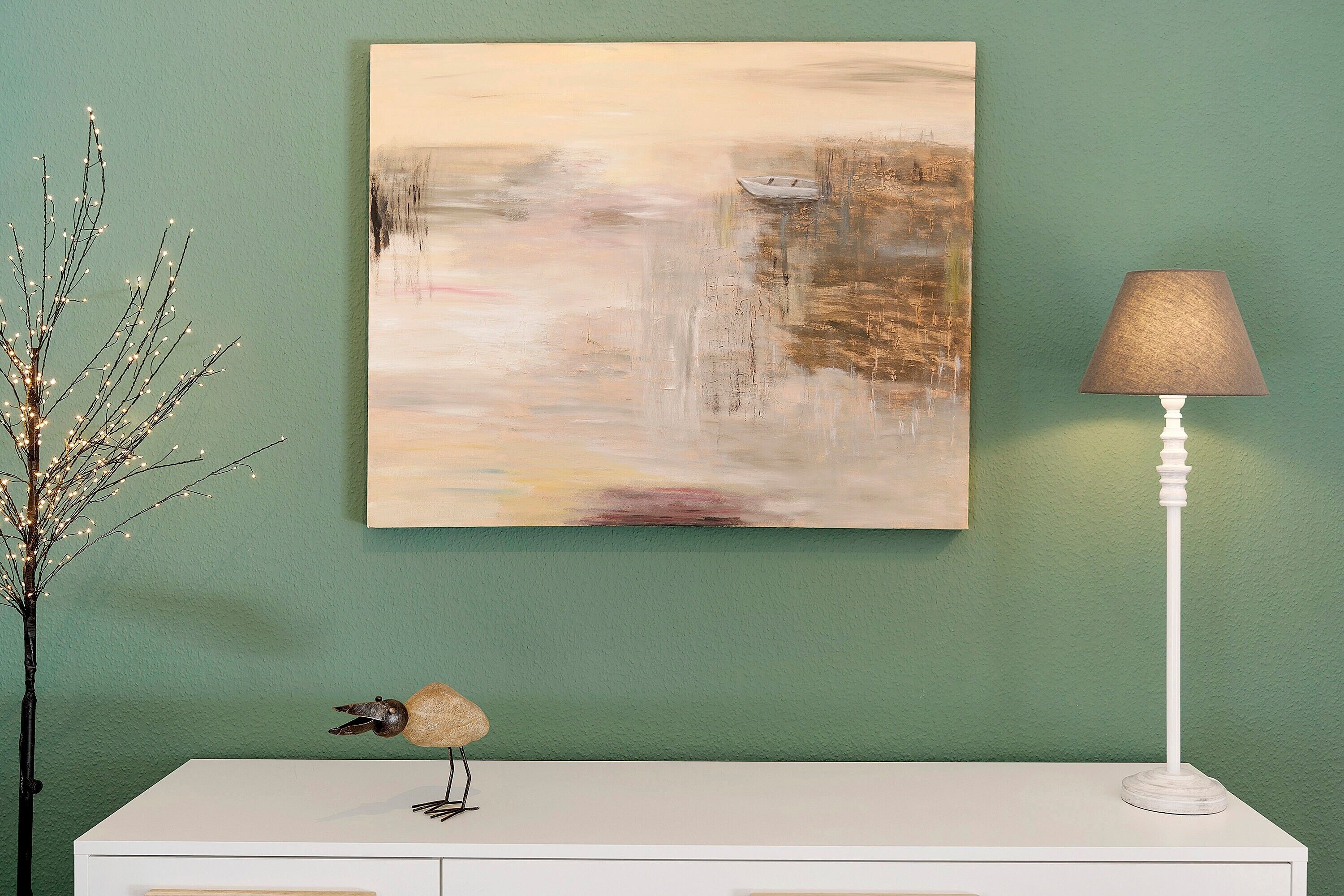 Un tableau, une lampe et une branche décorative posés sur une surface blanche, le tout sur fond de mur vert.