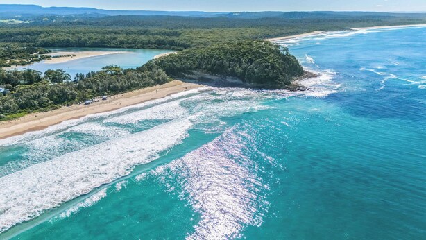 Eine Luftaufnahme einer Küstenlandschaft mit Strand, klarem türkisblauem Wasser und einer bewaldeten Insel.