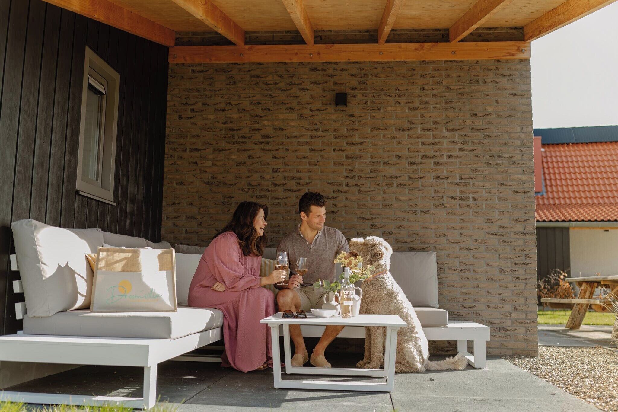 A couple sitting on a white sofa with a dog, a white table with drinks, and a stone wall in the background.