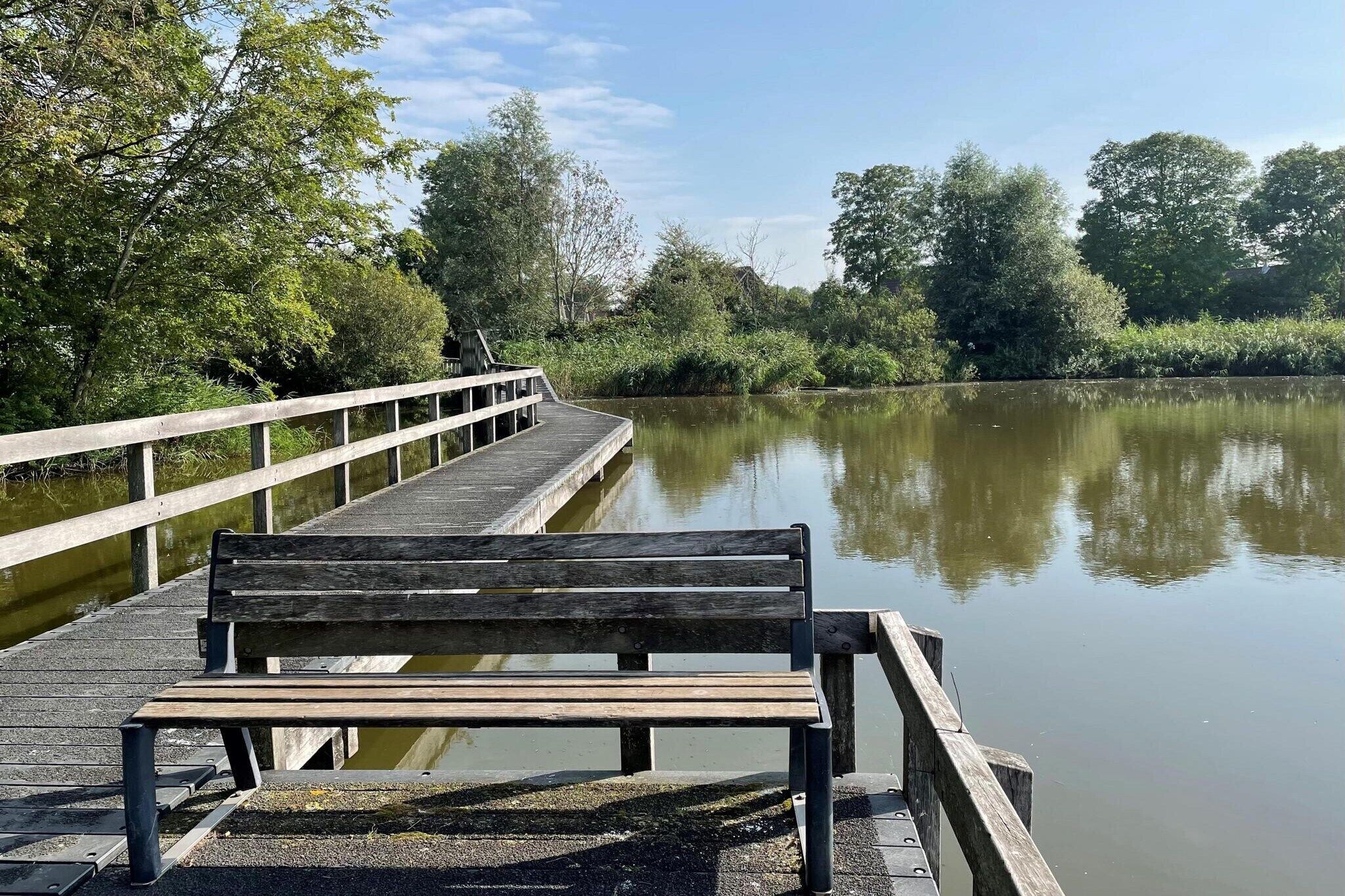 Un banc en bois, installé sur une jetée qui s’avance sur une étendue d’eau calme, entouré d’une végétation luxuriante.