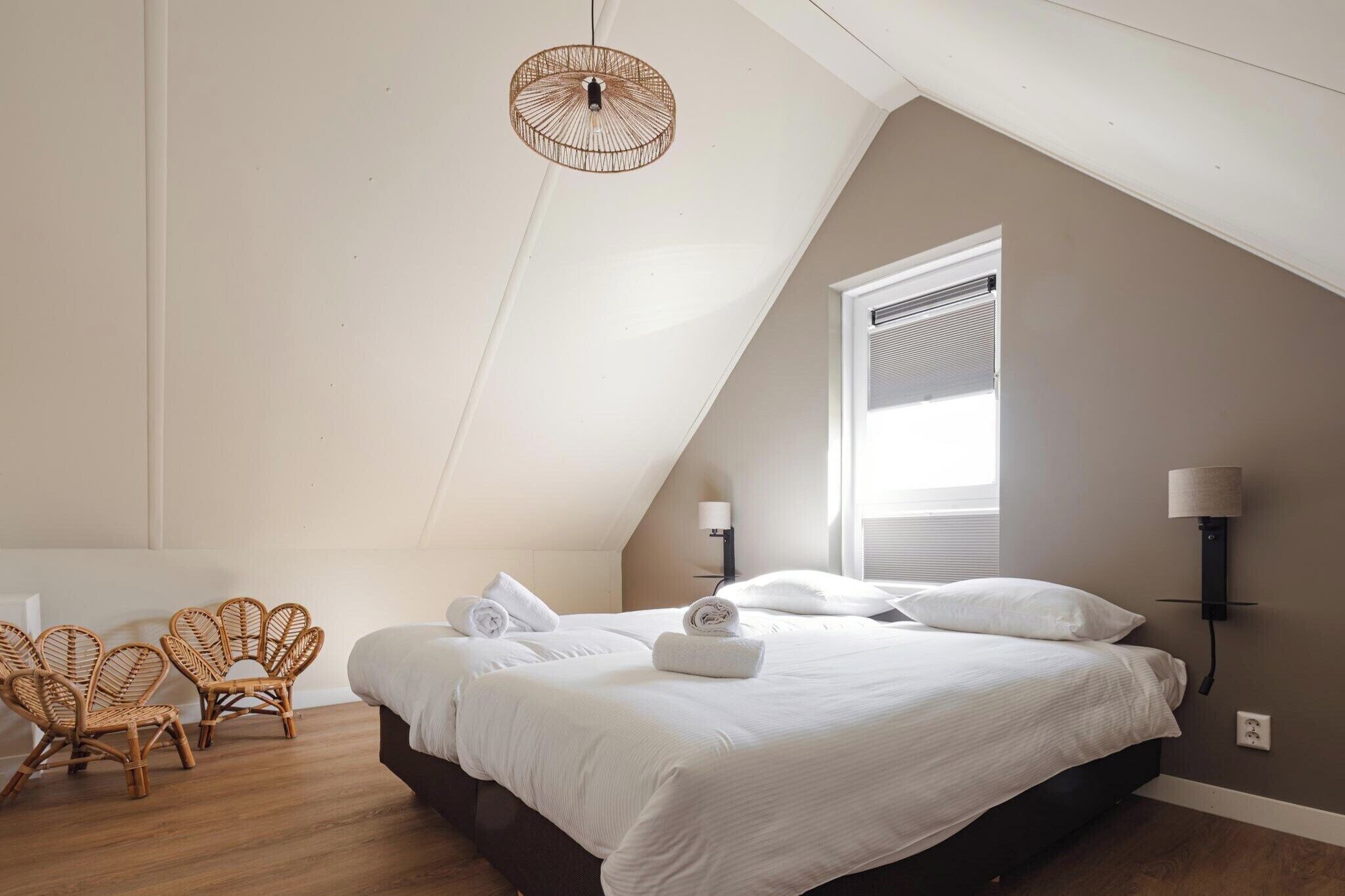 Quarto com uma cama grande, duas mesas de cabeceira, uma luminária pendente e duas cadeiras de vime.