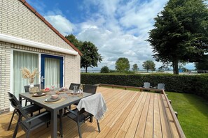 Een houten terras met een eettafel, een bakstenen gebouw met een blauwe deur en uitzicht op het landschap.