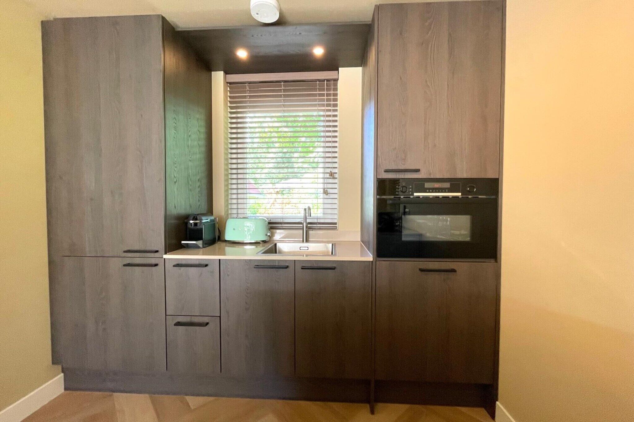 Una cocina compacta con armarios de madera oscura, horno empotrado y ventana con persianas.