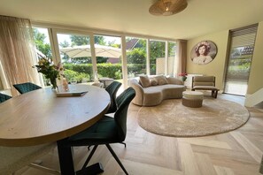 A spacious living area with a round wooden table, green chairs, a beige sofa, and a large window offering a view of the outdoors.