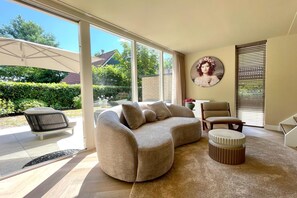 Ein modernes Wohnzimmer mit einem beigen Sofa, einem runden Ottomanen, einem Sessel und einem großen Fenster mit Blick auf einen Garten und ein Haus.