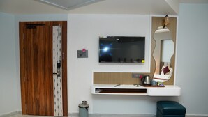 A room with a wooden door, a mounted TV, a vanity with a mirror, a small stool, and a trash can. - Hotel KAMA (Danapur)