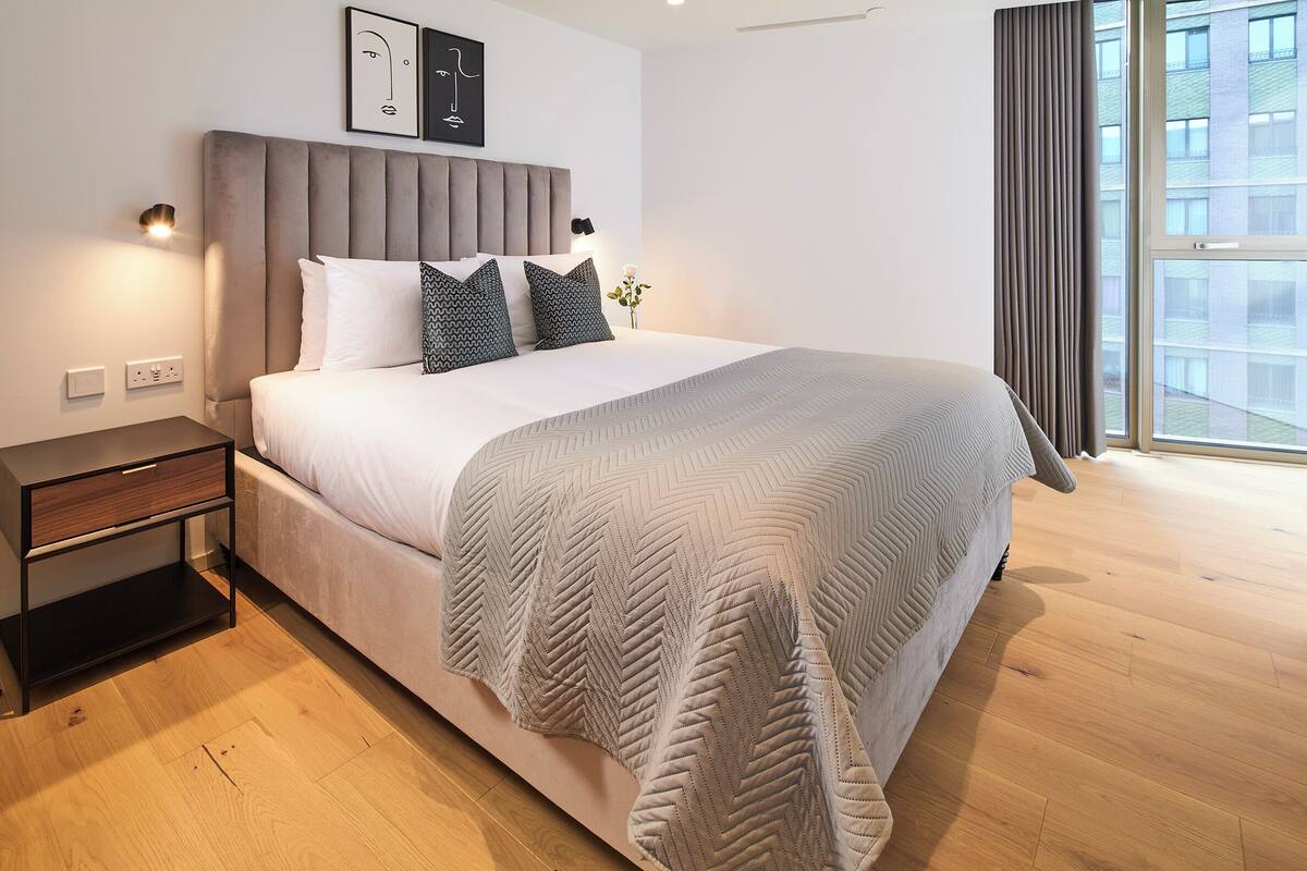 A modern bedroom with a large bed, a nightstand, a wall-mounted light, and a window with curtains.