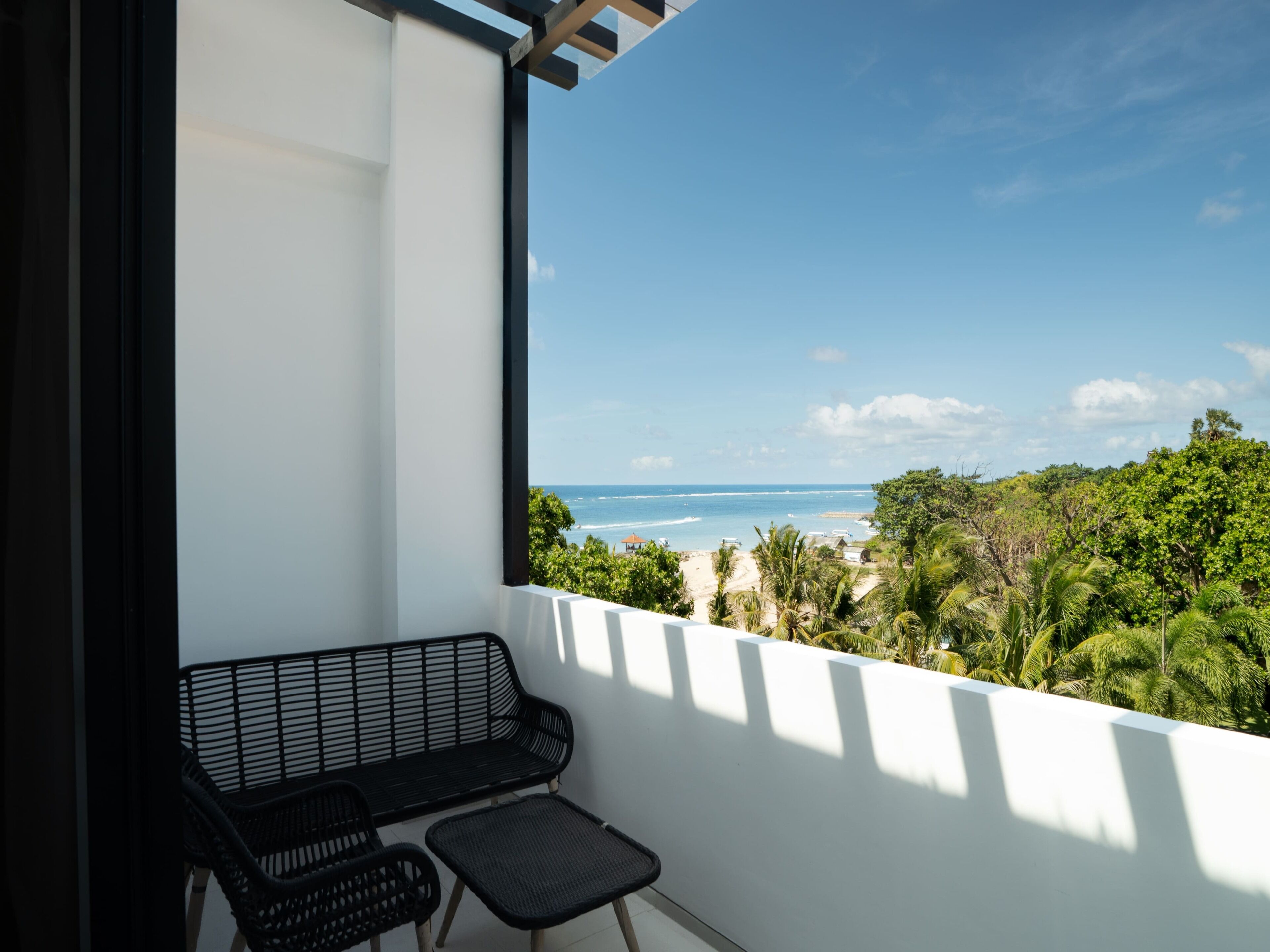 A balcony with a black metal bench and a small table, overlooking a beach and greenery.