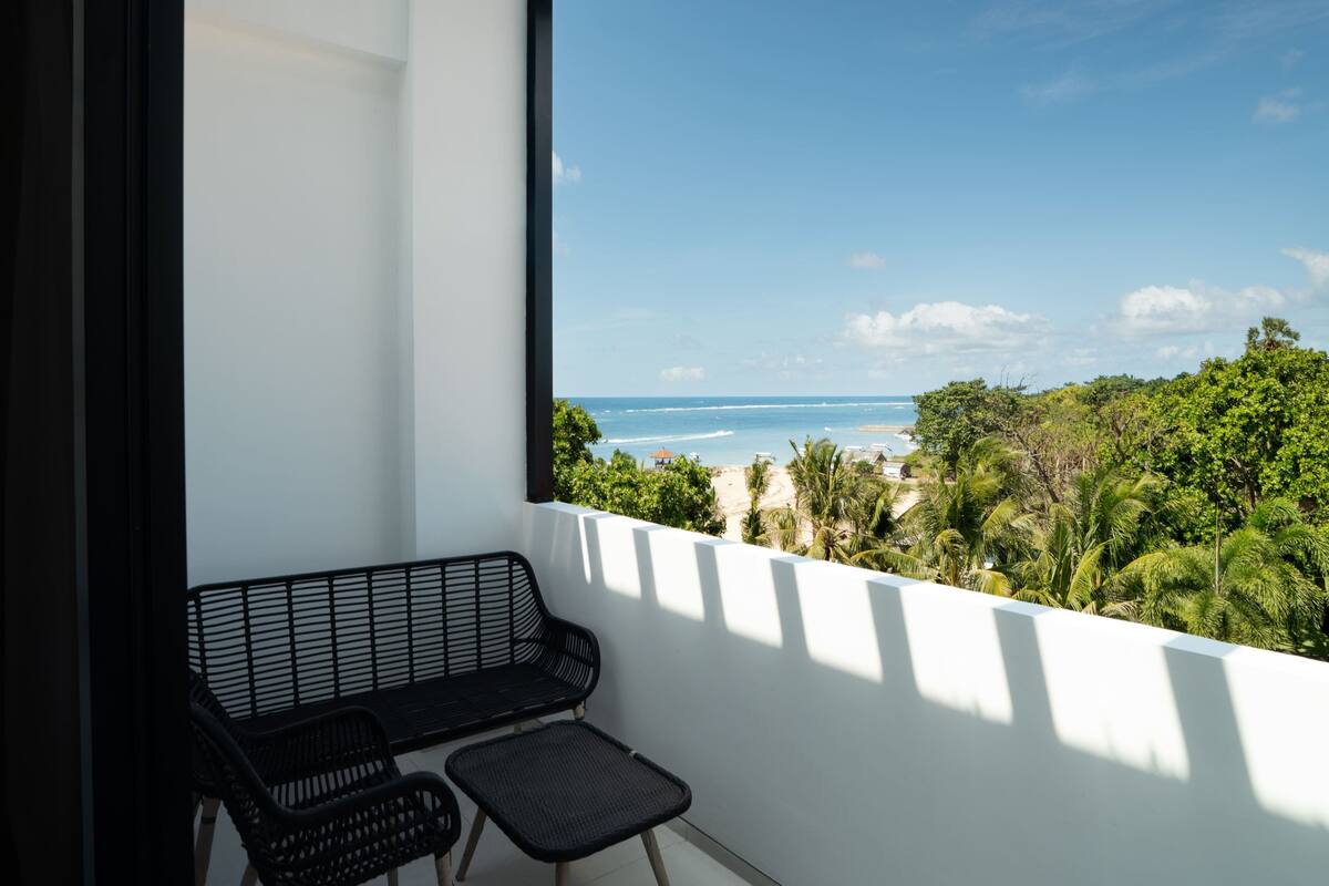 Ein Balkon mit einer schwarzen Metallbank und einem kleinen Tisch, mit Blick auf einen Strand und viel Grün.