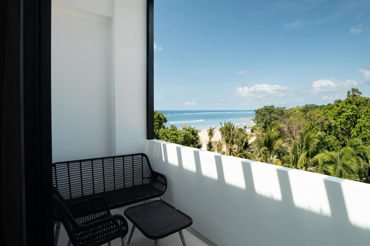 A balcony with a black metal bench and a small table, overlooking a beach and greenery.