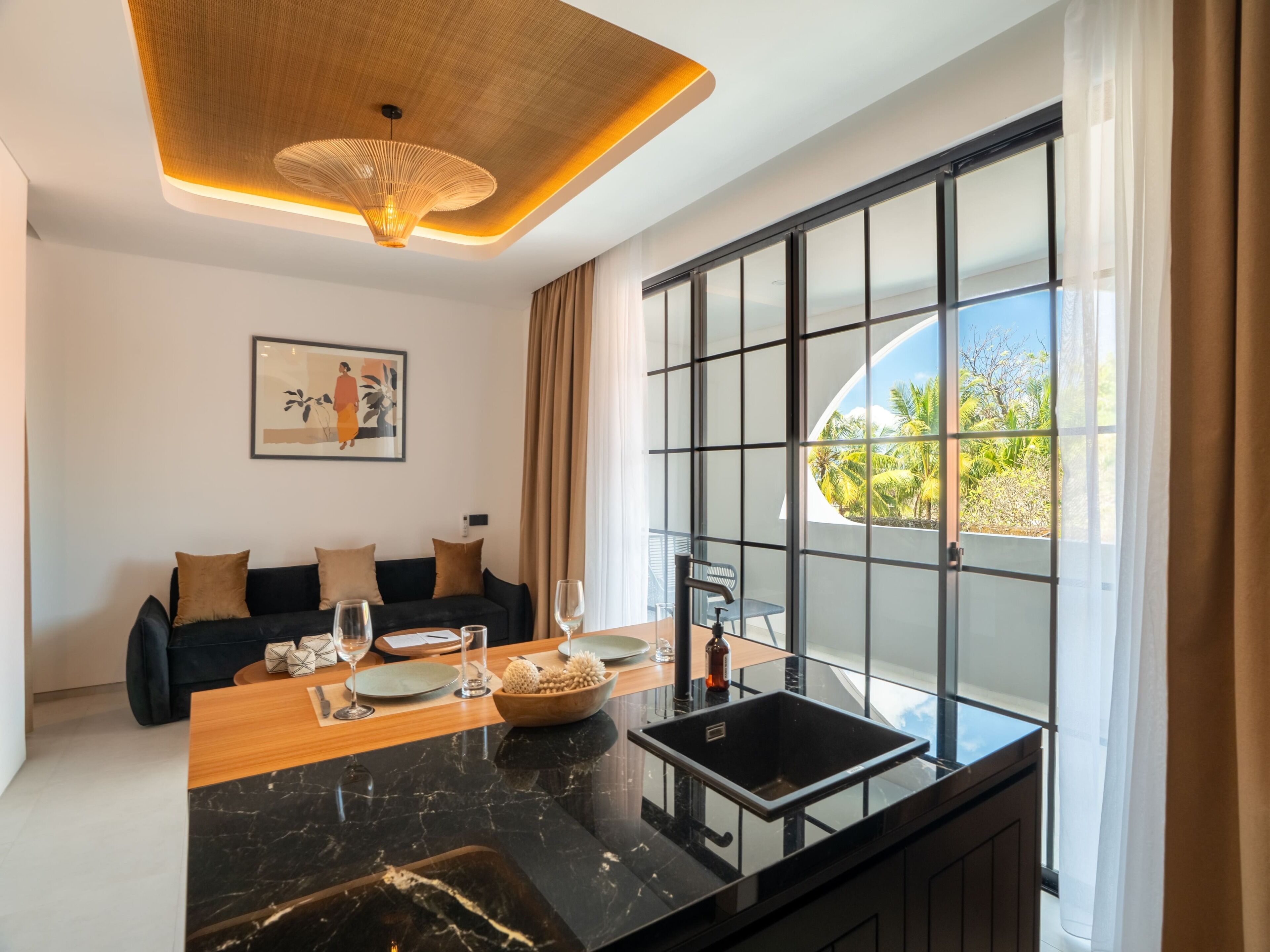A modern kitchen with a black marble countertop, a dining table with chairs, a sofa, and a large window with a view of greenery.