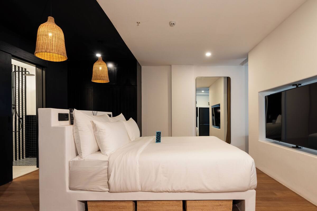 Une chambre moderne avec un lit blanc, deux lampes suspendues en osier, un grand miroir et un téléviseur fixé au mur.