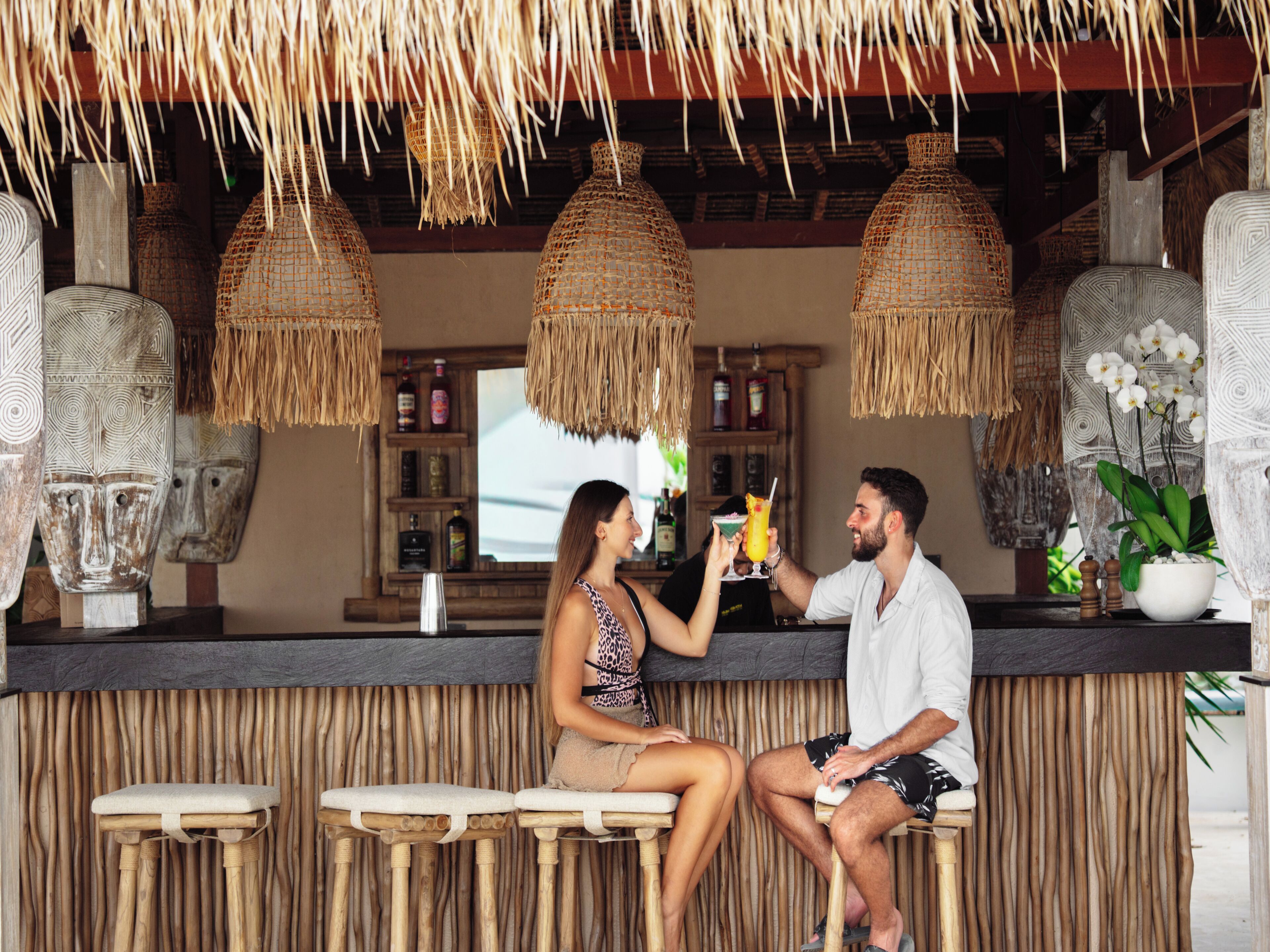 A bar area with woven ceiling decorations, wooden stools, and a counter with a display of bottles.