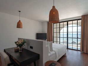 A modern hotel room with a bed, a TV, a dining table with a vase of flowers, and a view of the sea through large windows. - Kiara Beachtown Nusa Dua (Nusa Dua)