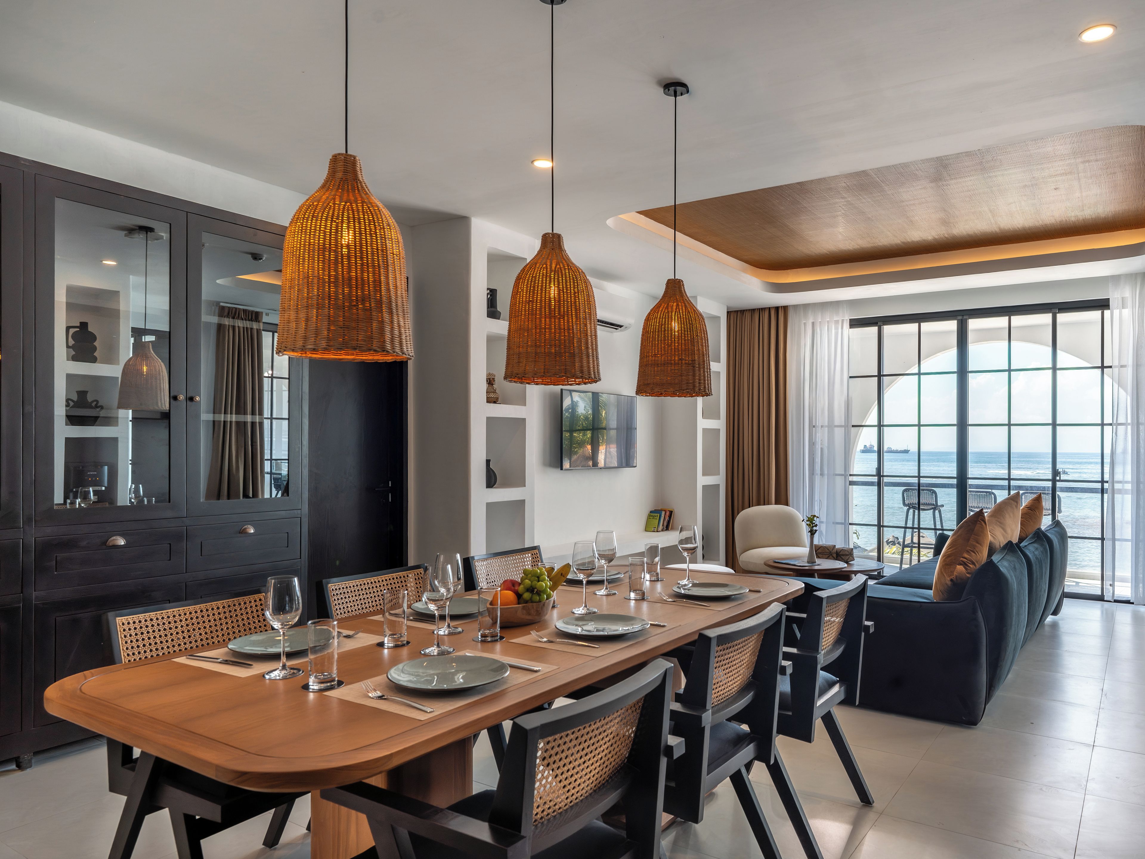 A modern dining area with a wooden table set for four, surrounded by wicker chairs, and a large window offering a view of the sea.