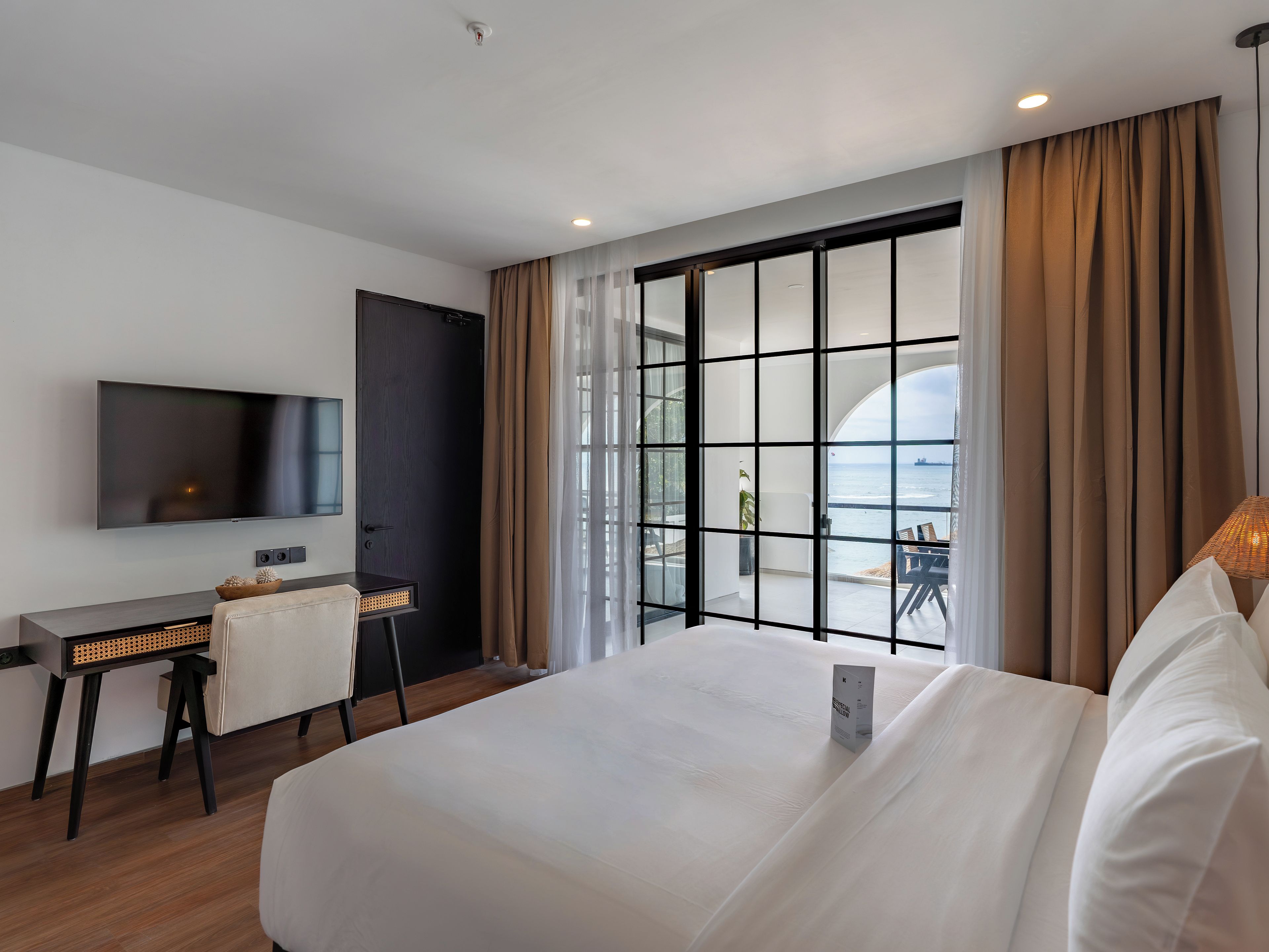Une chambre d’hôtel avec un grand lit, un bureau avec une chaise, une télévision et une vue sur la mer, visible à travers une porte vitrée.