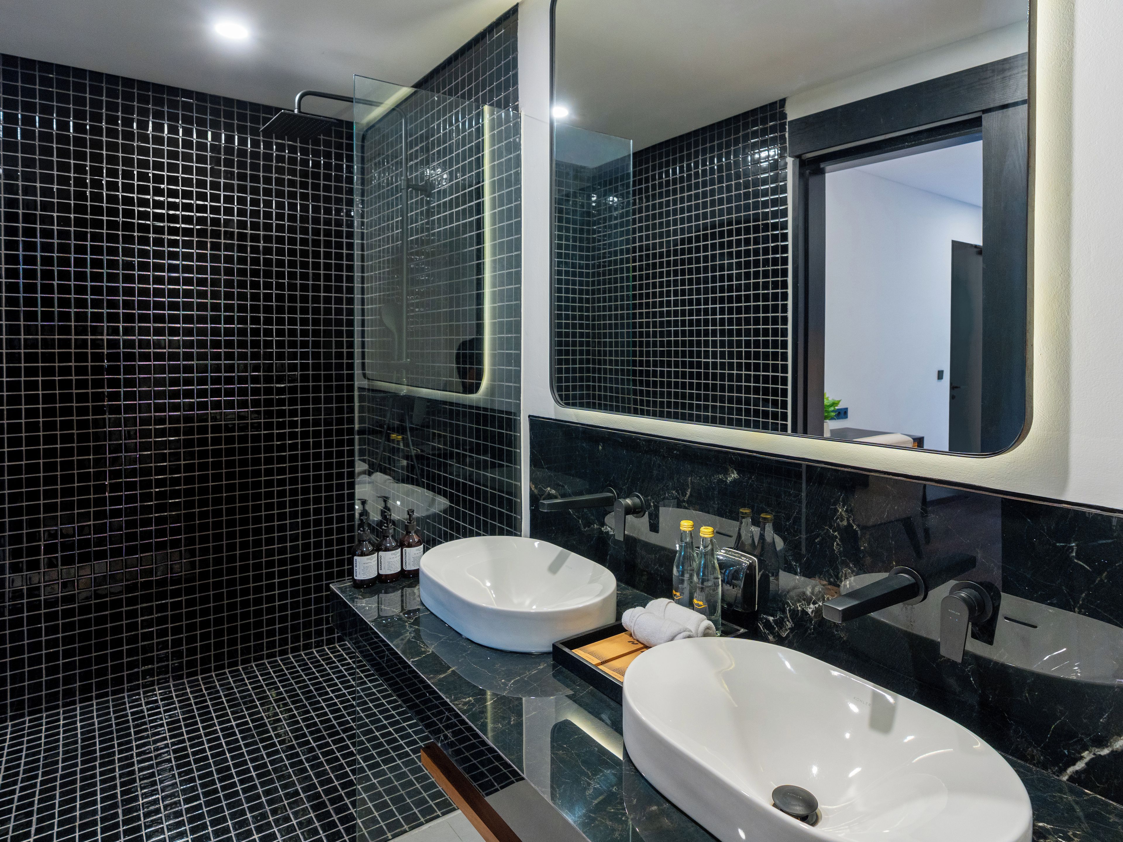 A modern bathroom with black tiled walls, a glass shower enclosure, and two sinks with faucets.