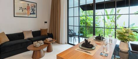 A modern living room with a sofa, coffee table, dining table, and large windows with a view of greenery.