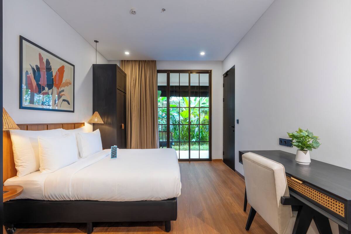 Ein modernes Hotelzimmer mit Bett, Schreibtisch, Stuhl und Blick ins Grüne durch das Fenster.