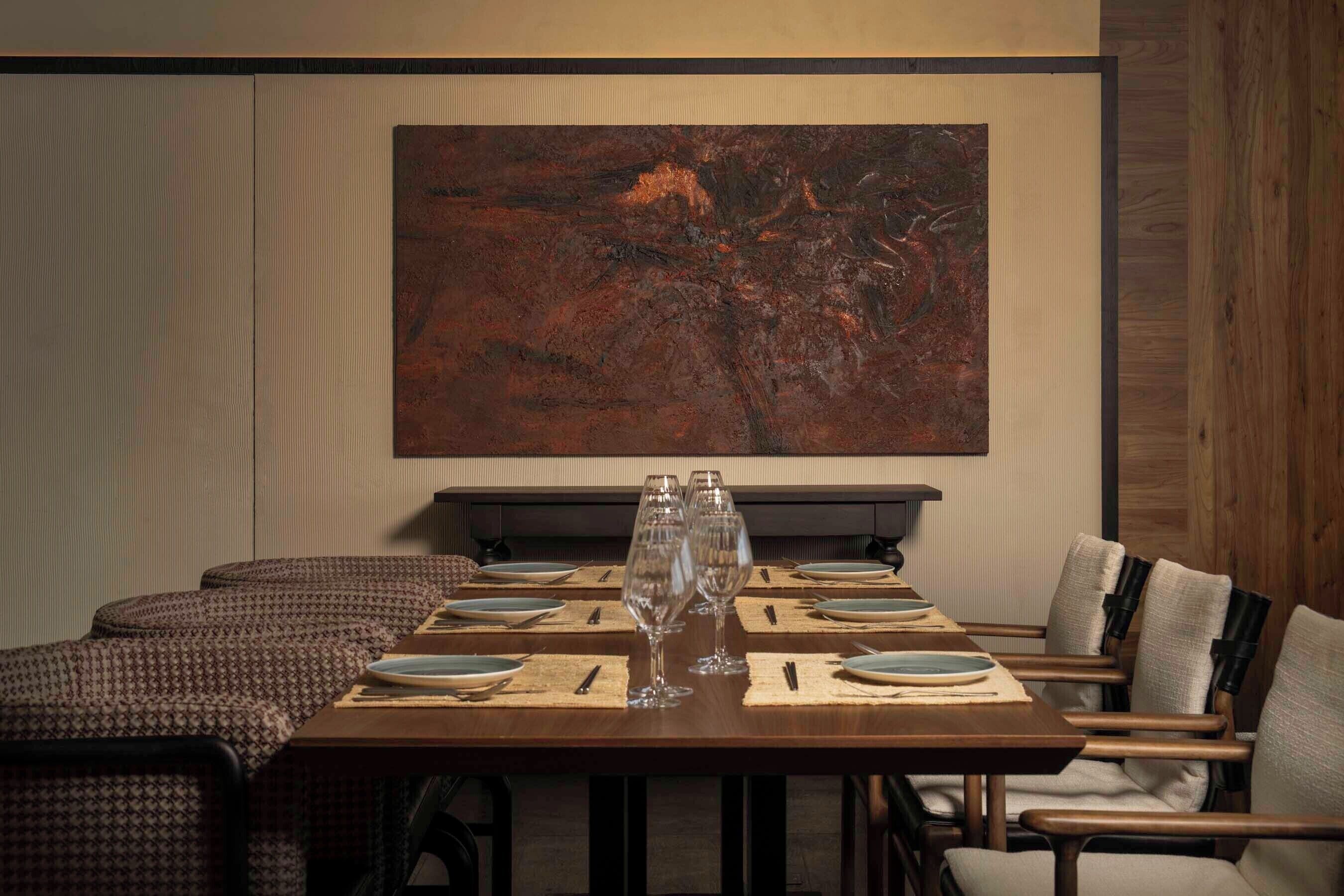 Un comedor con una mesa dispuesta para cuatro, con copas de vino y platos, y un cuadro abstracto grande sobre ella.