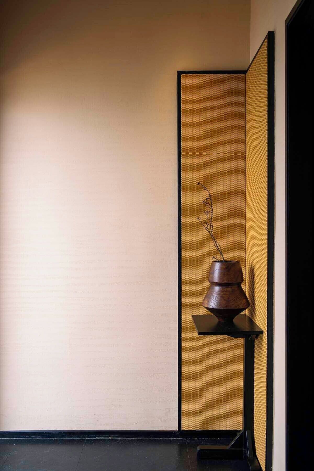 Una habitación con pared de esteras de tatami, un estante de madera con una maceta y suelo negro.