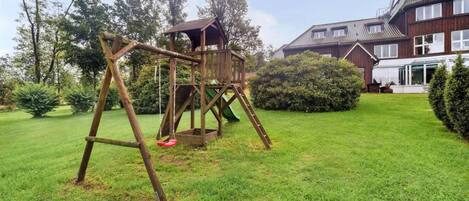 Un columpio de madera en un jardín con césped y una casa grande al fondo.