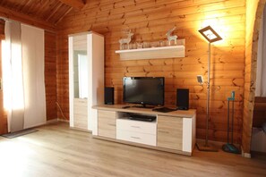 A wooden-paneled room with a TV on a white entertainment unit, a shelf with decorative items, and a standing lamp.