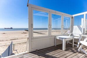 Un balcon donnant sur la plage, avec vue sur l’océan, une terrasse en bois et des meubles blancs.