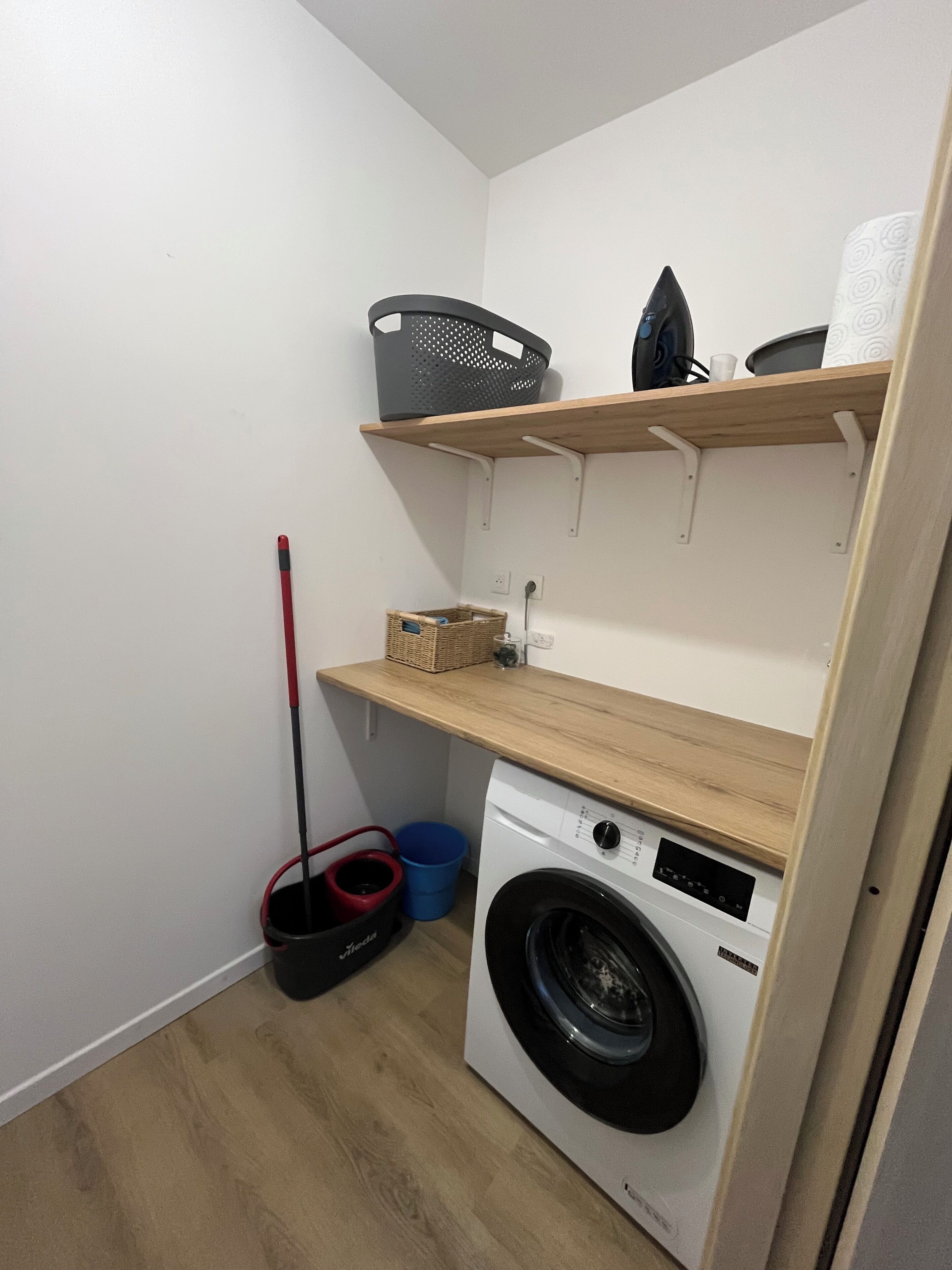 Une buanderie équipée d’un lave-linge, d’une serpillière, d’un balai et d’étagères contenant des paniers et une bouilloire.