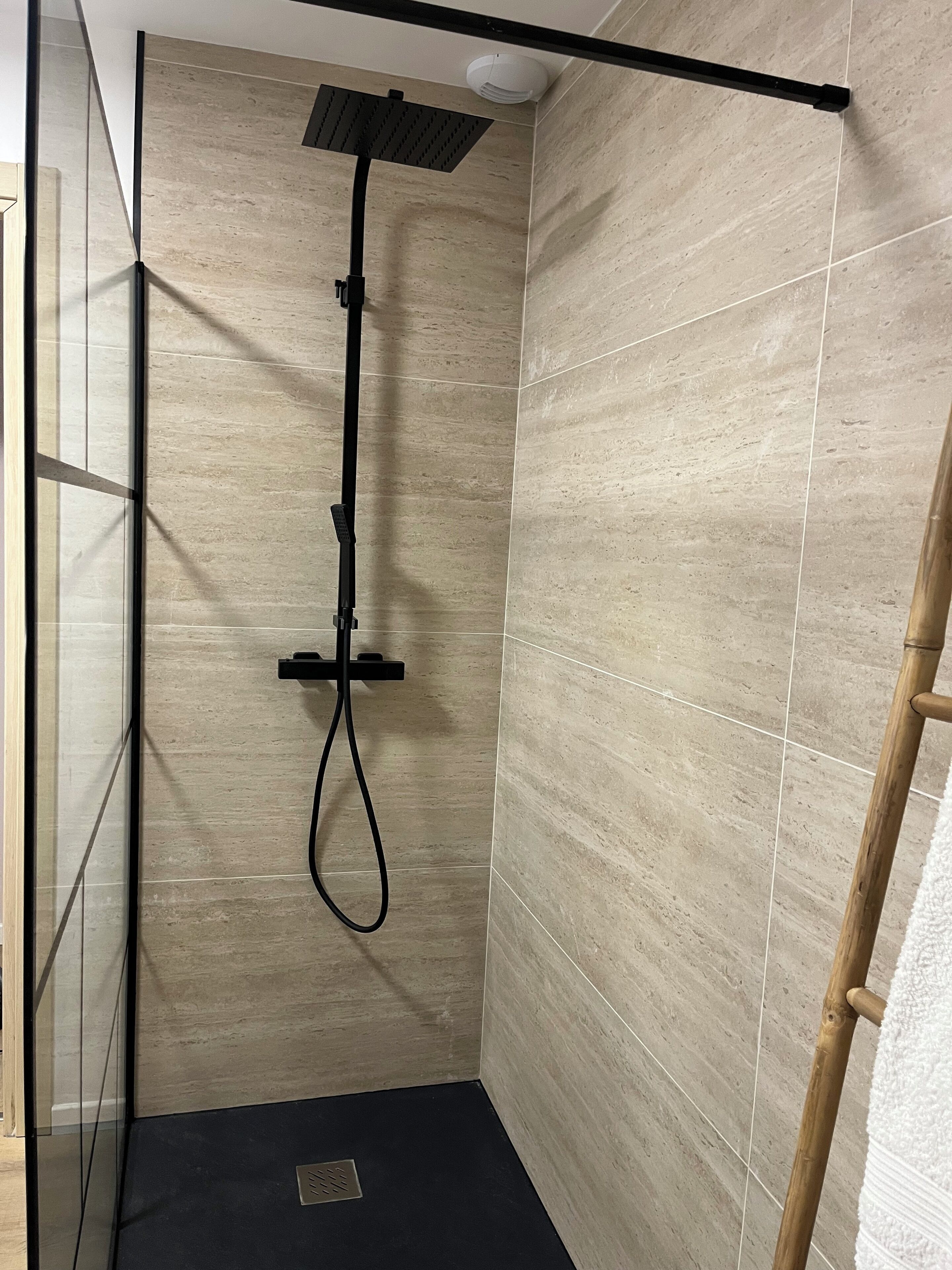 A shower with a black hose and a handheld showerhead, tiled walls, and a wooden towel rack.