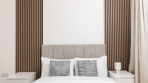 Ein Bett mit weißer Bettwäsche, zwei Kissen, ein Nachttisch mit Lampe und ein Kopfteil aus Holzleisten.