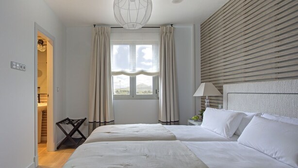 Habitación de hotel con dos camas, una ventana con cortinas, una lámpara colgante y una mesita de noche con lámpara.