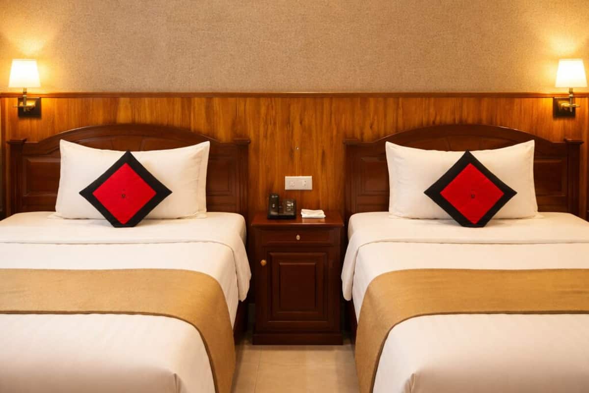 A hotel room with two beds, wooden headboards, a nightstand with a lamp, and a wall-mounted light fixture.
