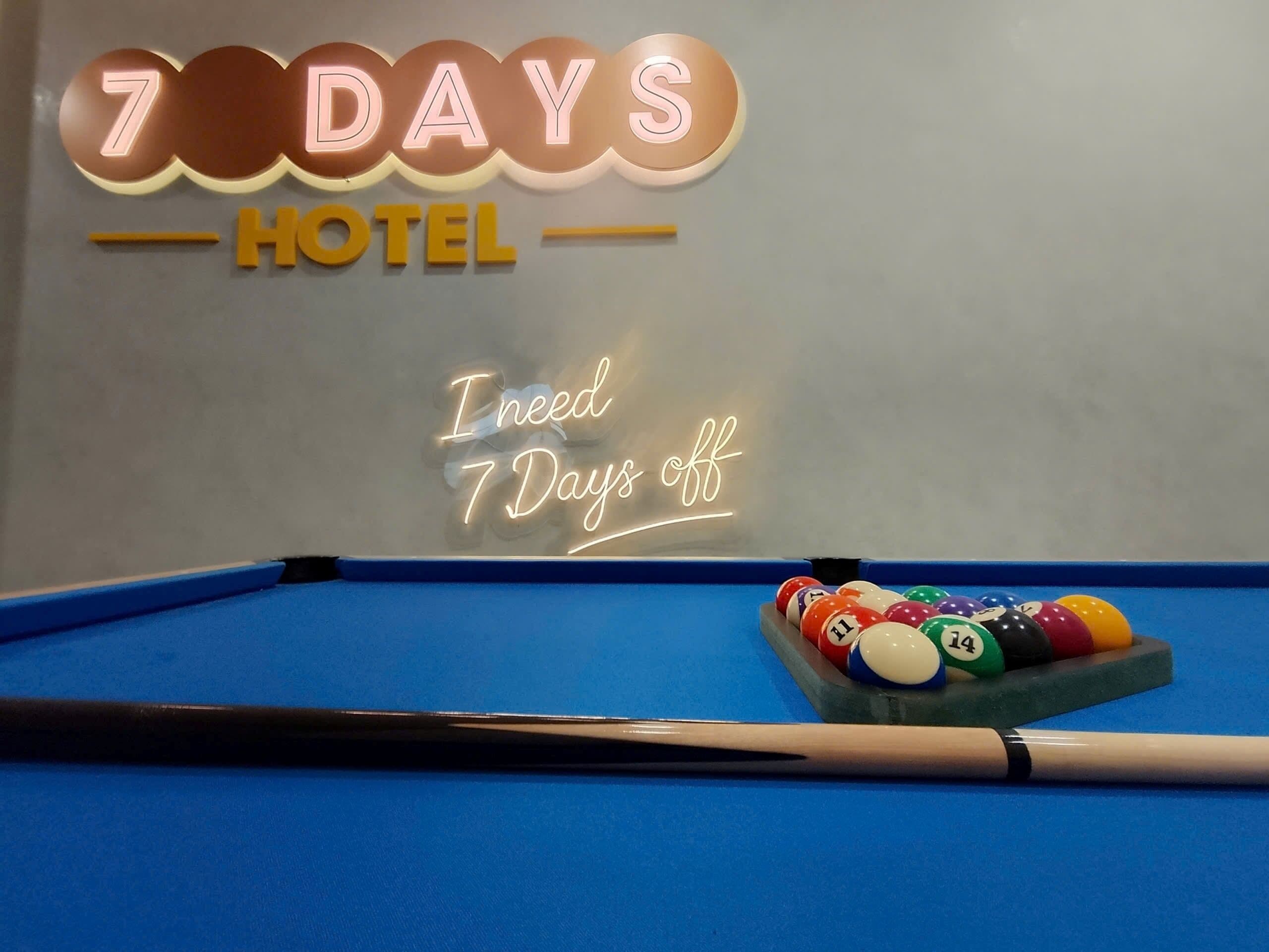 Una mesa de billar con taco y bolas, un letrero de neón para '7 DAYS HOTEL', y un cartel que dice 'Necesito 7 días libres'.