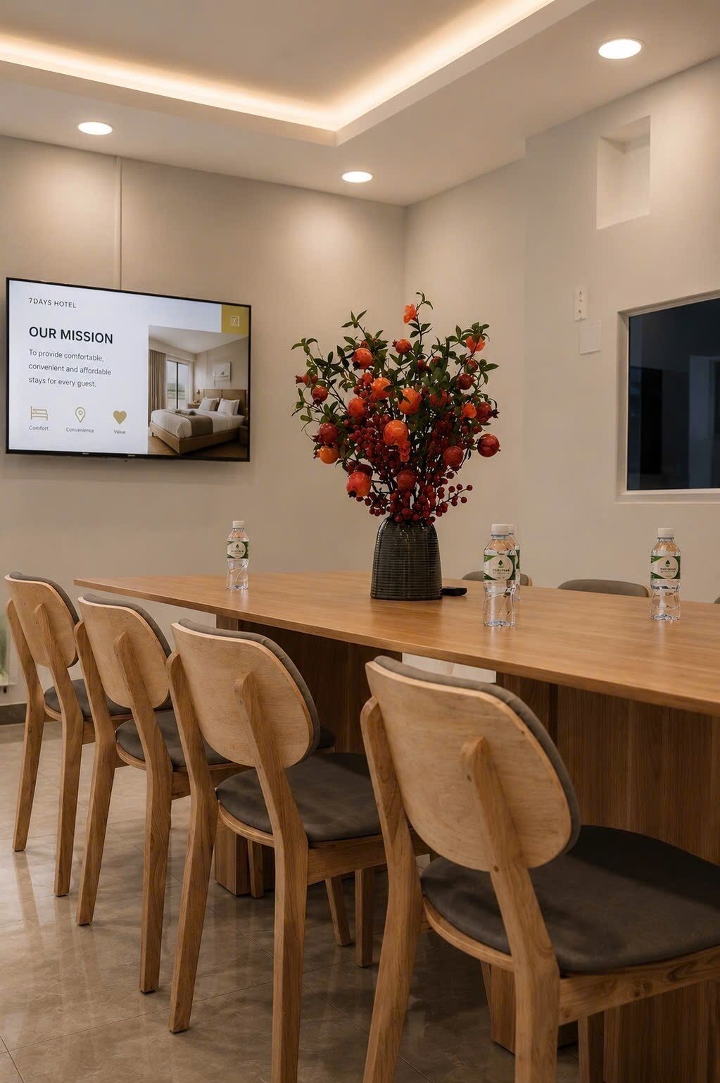 A conference room with a long wooden table, four chairs, a potted plant, and a screen displaying hotel information.