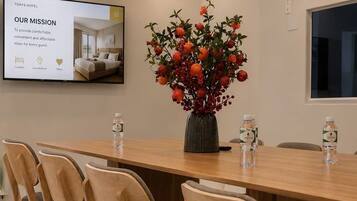 A conference room with a long wooden table, four chairs, a potted plant, and a screen displaying hotel information.