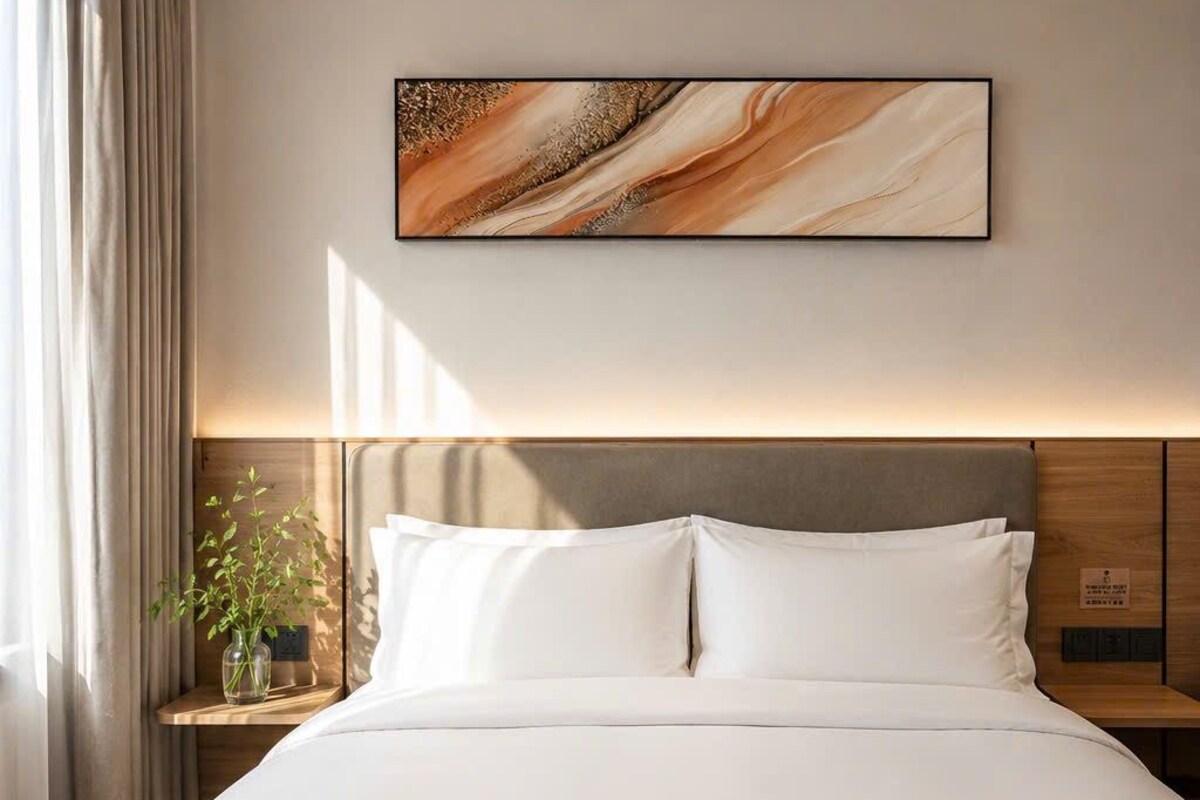A modern hotel room with a large bed, a nightstand with a lamp, a wall-mounted artwork, and a window with curtains.