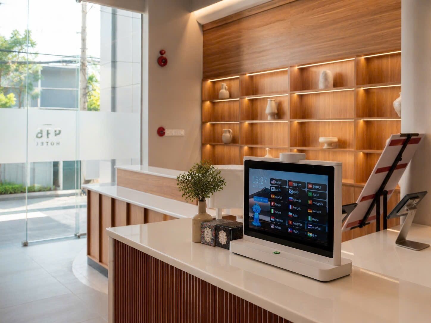 Un mostrador de recepción de hotel con una pantalla digital que muestra información de habitaciones, una planta pequeña y un cartel.