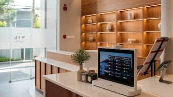 Un mostrador de recepción de hotel con una pantalla digital que muestra información de habitaciones, una planta pequeña y un cartel.