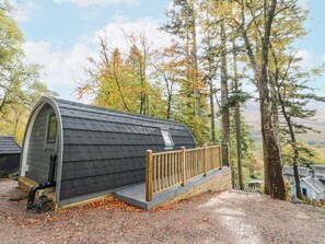 A small, curved wooden cabin with a wooden deck and a metal railing, surrounded by trees and a clear sky. - Elleric Pods - The Last Resort (Appin)