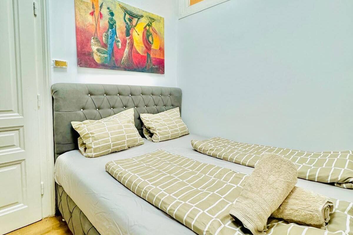 A bedroom with a bed, a grey upholstered headboard, a painting on the wall, and a window with frosted glass.