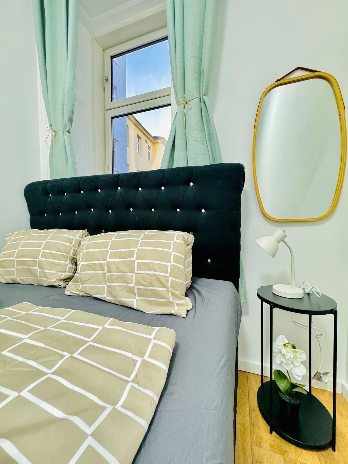 A bedroom with a tufted headboard, two patterned pillows, a bedside table with a lamp, and a mirror.