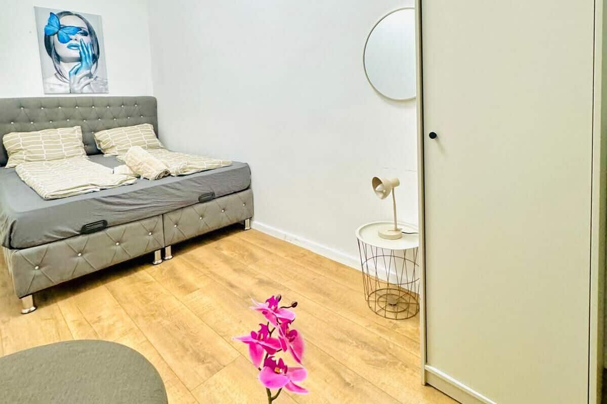 A modern bedroom with a grey sofa, a bed with patterned pillows, a wall-mounted mirror, and a potted orchid.