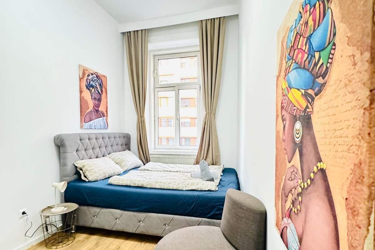 A bedroom with a bed, a sofa, a small table with a plant, a remote control, and a painting on the wall.