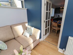 A living room with a beige sofa, a dining table with chairs, and a kitchen area visible through an open door. - Linn Cottage, Bellingham (Hexham)