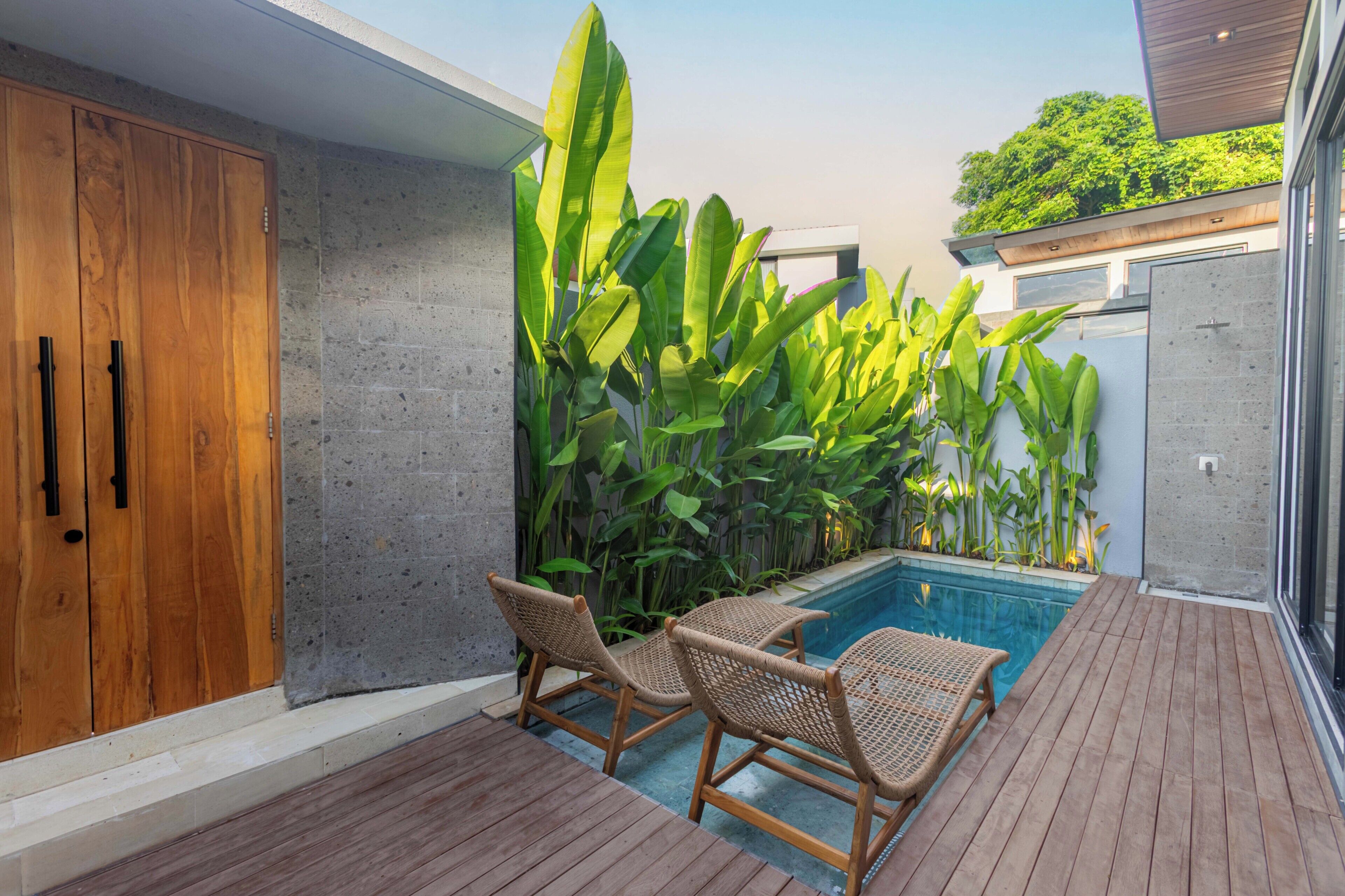 A modern outdoor pool area with wooden deck, wicker chairs, and lush greenery.